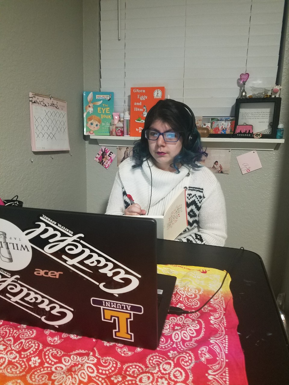 Fermina Lopez a young Latina woman sitting at a table in her home, looking at her laptop that is covered in stickers. She is wearing a big cozy white sweater and listening to headphones plugged into the computer.