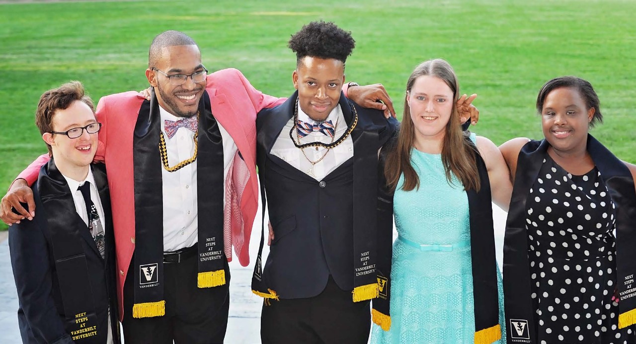 Next Steps at Vanderbilt graduates Next Steps at Vanderbilt and shows 5 students on their graduation day, dressed in formal clothes and wearing academic sashes with the Vanderbilt logo around their necks. There are three young Black men, one young white woman, one young Black woman and one young white man. Two of the grads have Down syndrome.