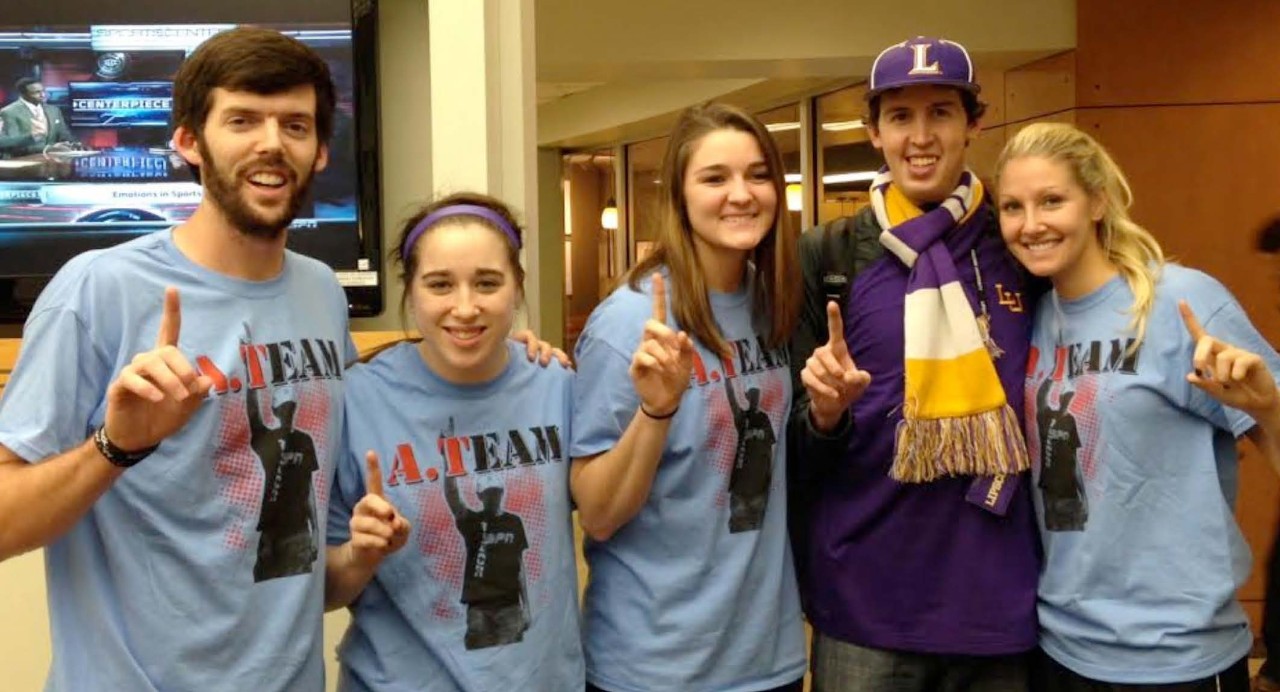 Lipscomb University IDEAL students Lipscomb IDEAL program and shows 5 white college students, two boys and three girls with and without intellectual disabilities. 4 are in matching T-shirts that say “A-Team” and the other young man is wearing Lipscomb sports colors of purple and gold. They are all holding up 1 finger in a kind of salute and smiling