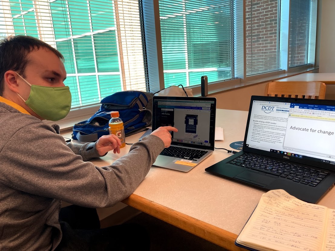 Carter Poland Carter Poland, a young white man wearing a green face mask, seated at a table with a laptop in front of him, working