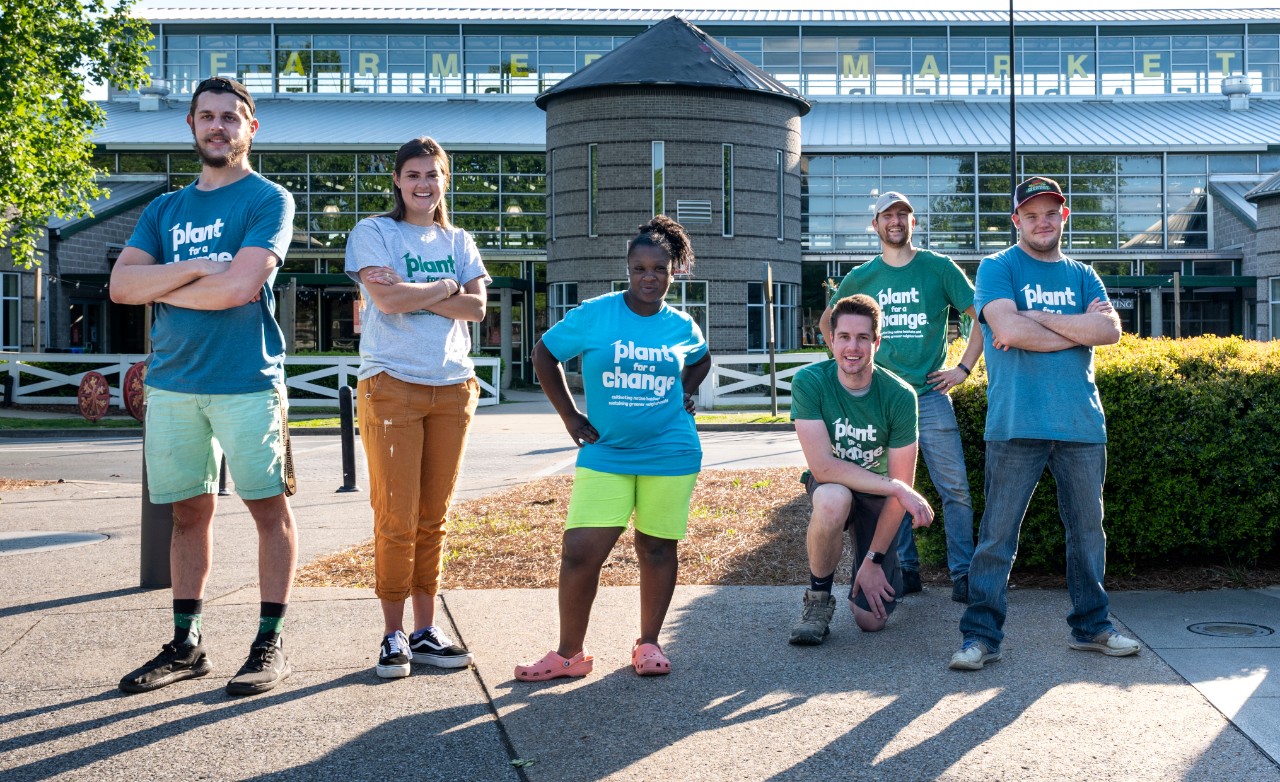 Plant for a Change crew group photo of people with and without disabilities – two young women, and four young men. They are posed standing and kneeling outside of the Nashville Farmer’s Market, where Plant for a Change hosts a plant selling stand for their business. They are all wearing matching T-shirts that say “Plant for a Change.”