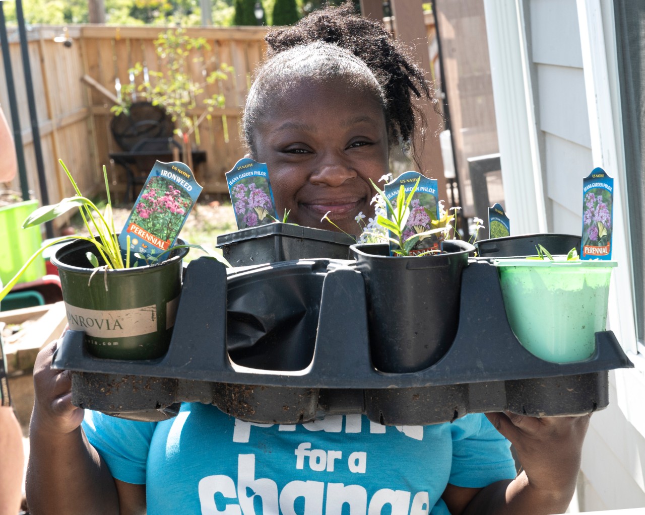 04-19-21 Plant for Change a young Black woman smiling very big, standing outside on a sunny day, and wearing a blue t-shirt that says “Plant for a Change.” She is holding up a tray of small potted plants just starting to sprout, with signs showing which plants or flowers they are, and smiling over the tray at the camera.
