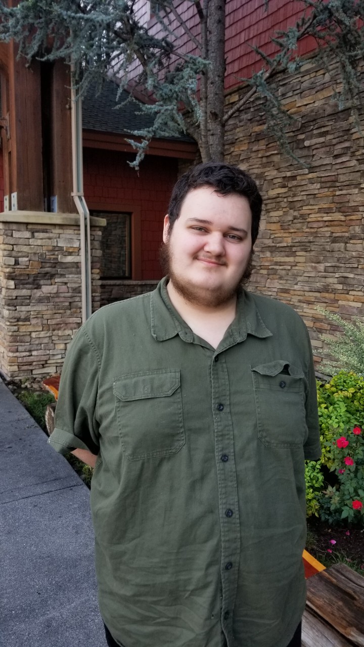 Mikey Newton a casual headshot of Mikey Newton, a young adult white man in a dark green button down short-sleeved shirt, with brown hair and a brown beard. He is standing with his hands behind his back and smiling