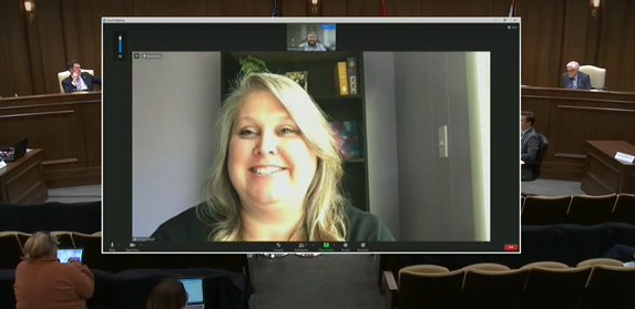 Council member Chrissy Hood – a white woman with blond hair, appears on a virtual meeting screen. Her virtual testimony is being heard in a legislative hearing room, with state legislators at the dark wood desk on the elevated dais