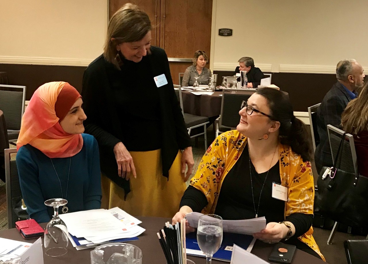 A photo of Wanda standing between two women talking to them as they sit at a table in a meeting room