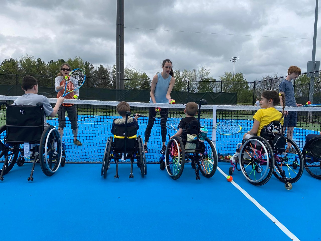 Wheelchair tennis at ABLE Youth's Super Sports Saturdays 4 kids and young adults are seated in wheelchairs in front of a tennis net on a tennis court holding rackets, facing a couple of instructors
