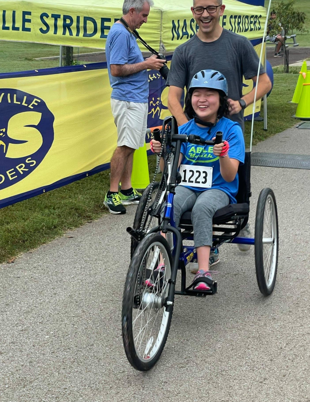 Hope & Possibility (R) Race a girl is seated on a large accessible tricycle that is low to the ground and an adult is running along behind her, helping push during the race