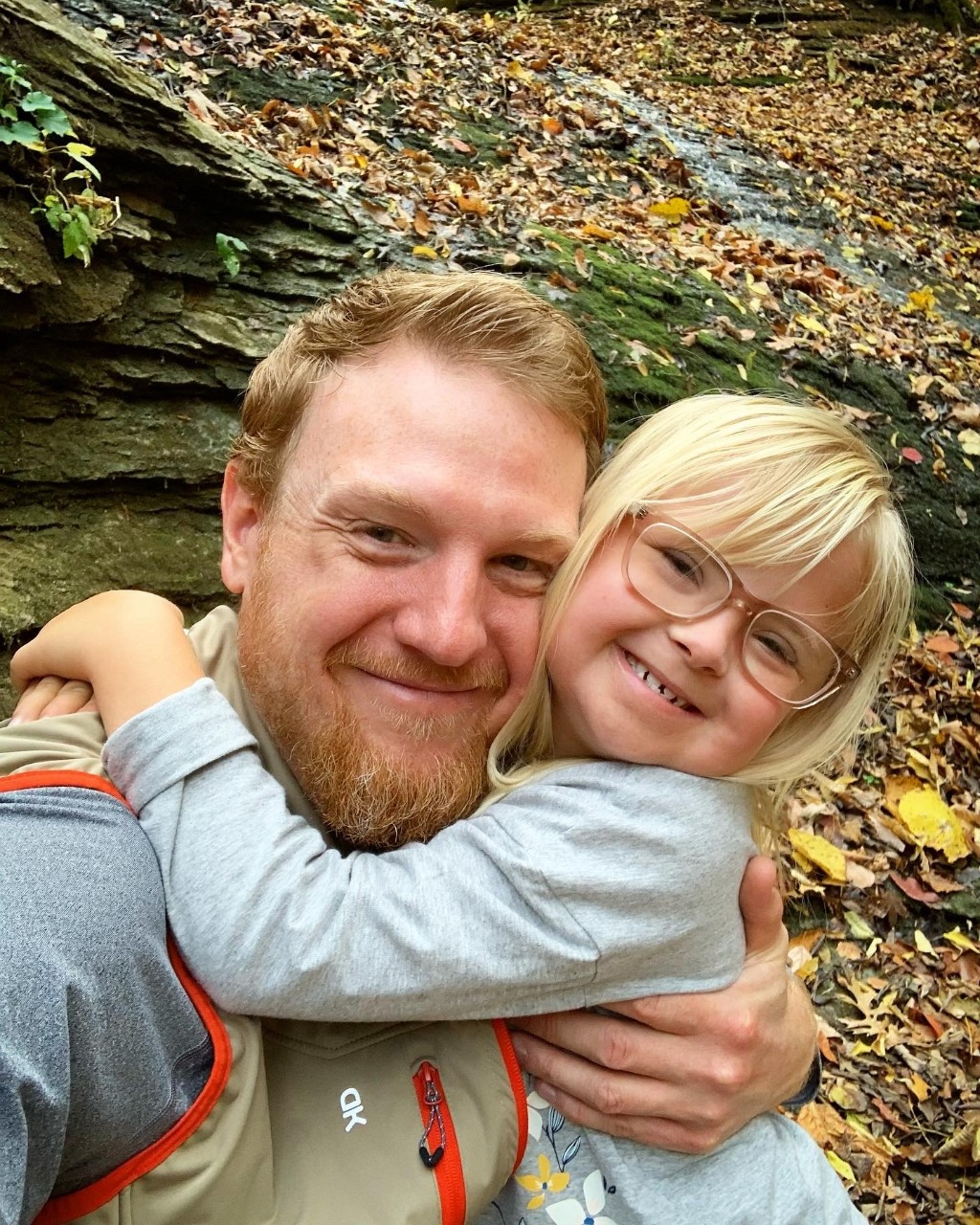 Jon is a young dad with reddish short hair and a beard and Lina is a young girl with light blonde hair and glasses who is tightly hugging her dad’s neck with a big grin on her face, fall leaves in the forest behind them