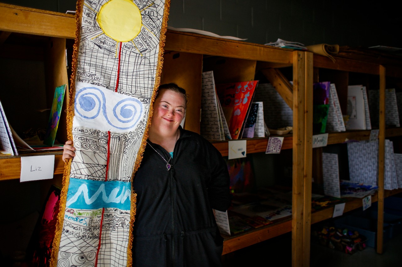 Katie Hamilton posing with her art a young white woman with down syndrome stands in front of some wooden shelves holding on to a large 3-D textile work of art that appears to include fabric and drawings showing the sun and other natural shapes