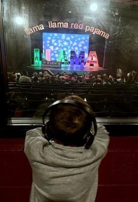 Child watches a recent NCT production from the Sensory Room photo at the Nashville Children's Theater with "Llama Llama Red Pajama" set design on stage, with the a child wearing noise canceling headphones leaning forward and watching the play from their new sensory room