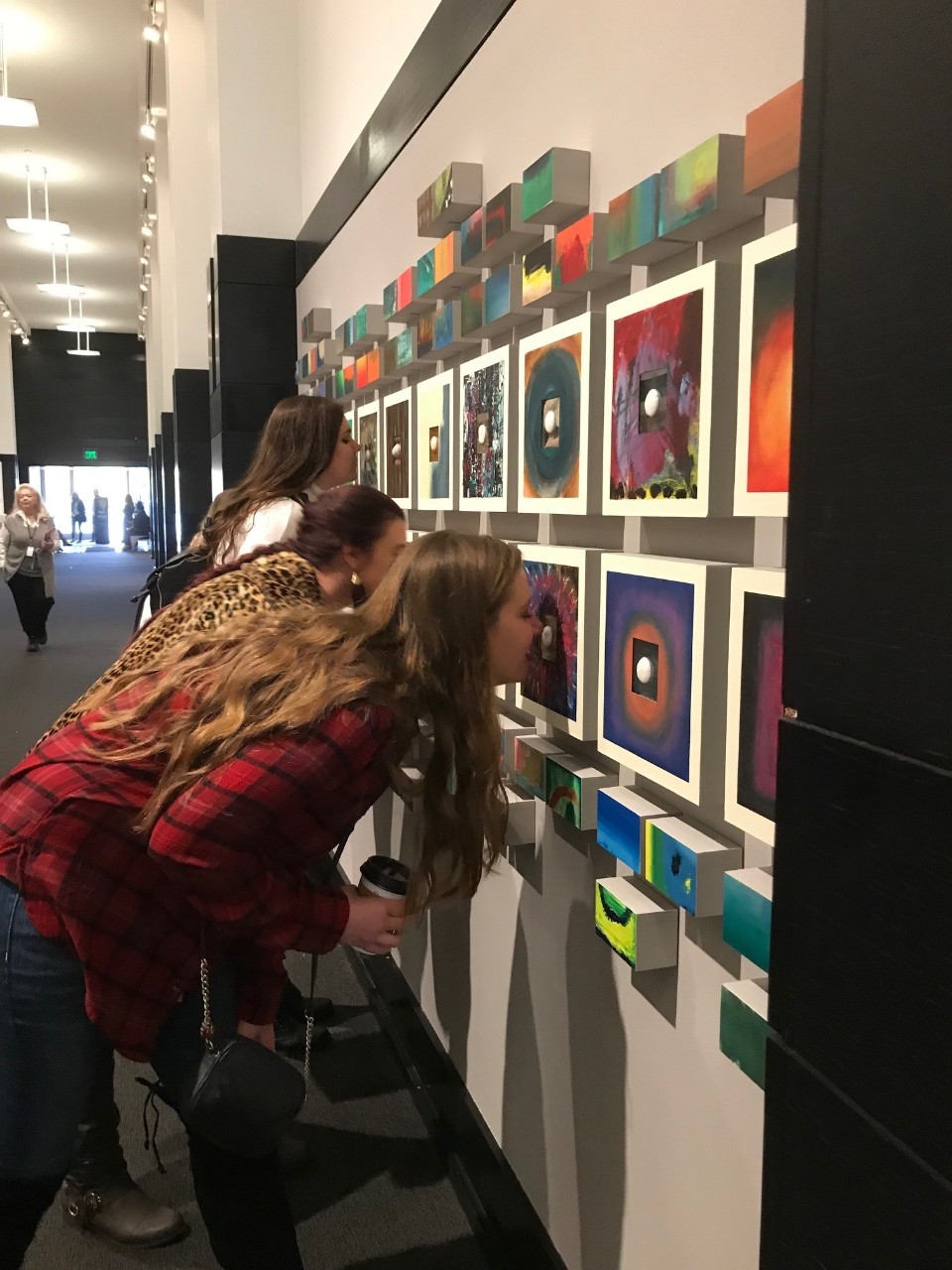 Multi sensory image several young women lean close to a display of several colorful textile squares mounted on a wall
