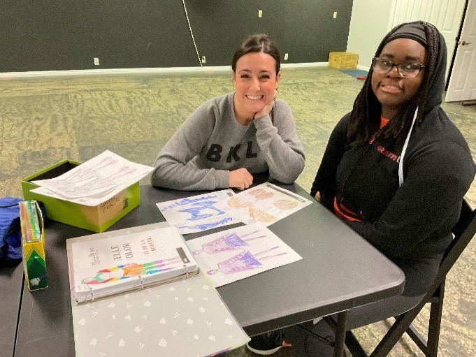 two women sit at a table together that is covered in colorful sketches of costumes and smile for the camera