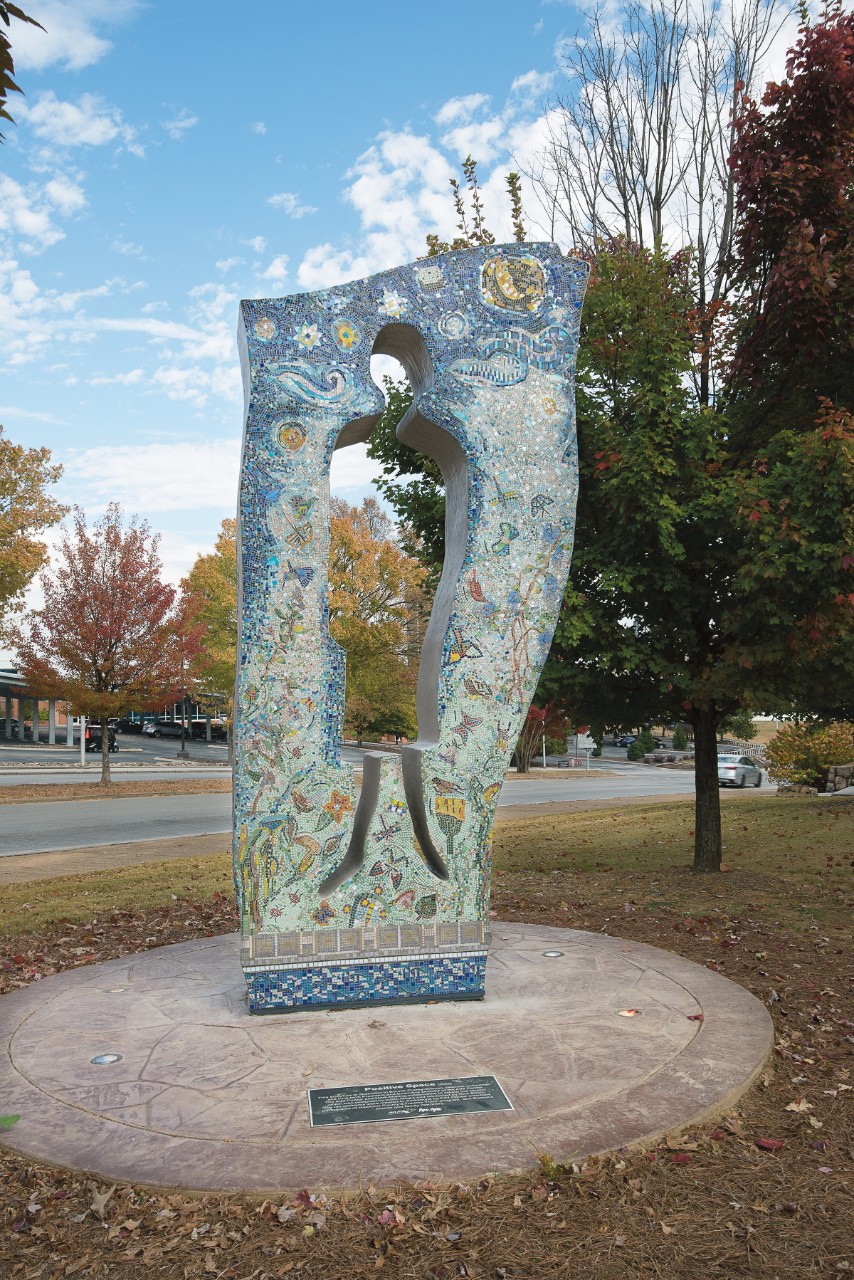 "Positive Space" sculpture a tall outdoor mosaic sculpture that surrounds an empty space of a human body – head, body, legs – and the surrounding mosaic tiles are in varying shades of blue and many other colors in the shapes of suns, moons, stars, butterflies, dragonflies, and similar figures