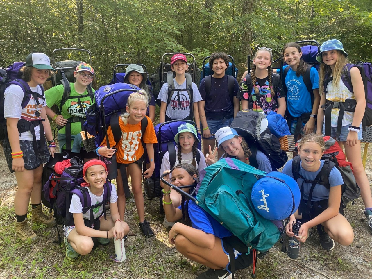 SYC Lumber Janes a photo of a diverse group of about 15 young girls in shorts, T-shirts and camping gear like boots and backpacks posing together in the woods and smiling