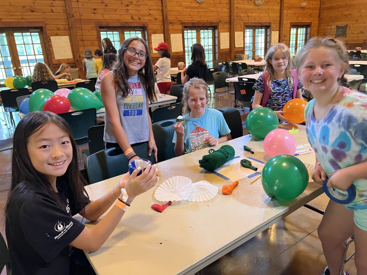 HOL Mischief a group of 5 young girls sit at a table in a large cabin with markers and balloons and smile for the camera