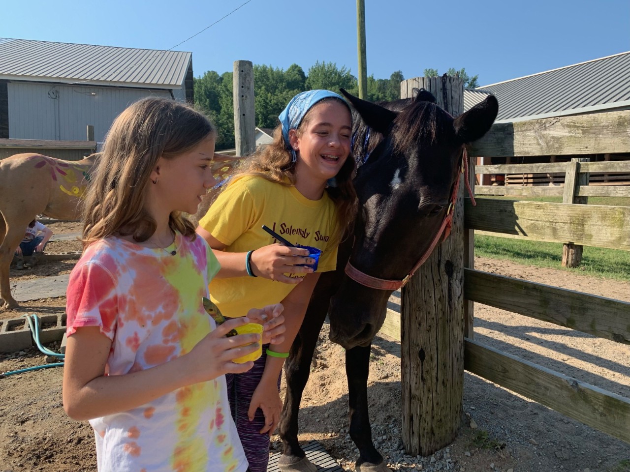 SYC Painting Horses two young girls lean near a black horse’s head at an outdoor fence while laughing; there’s a brown horse in the background with painted handprints on its side