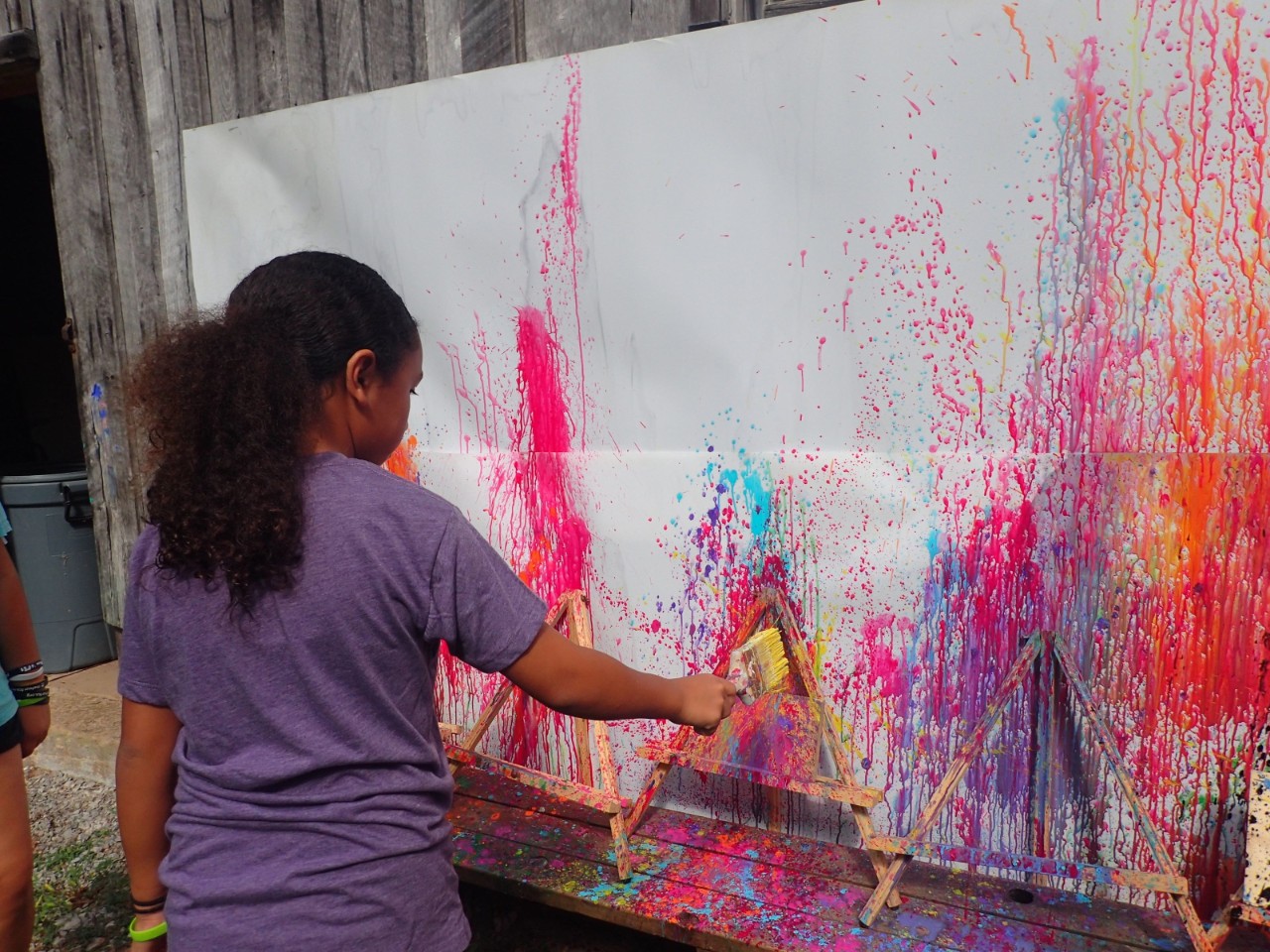 SYC Splatter Paint a photo of a young black girl with a purple shirt and a long ponytail holding a paintbrush in front of a huge piece of paper on a wall splattered with bright paint in pinks, oranges, reds and purples