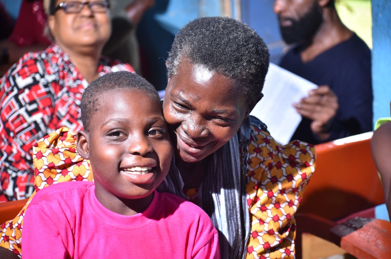 a young child in a bright pink shirt and an adult female family member with a colorful dress is shown leaning over his shoulder - the boy is looking at the camera directly and smiling, his family member is smiling down past him