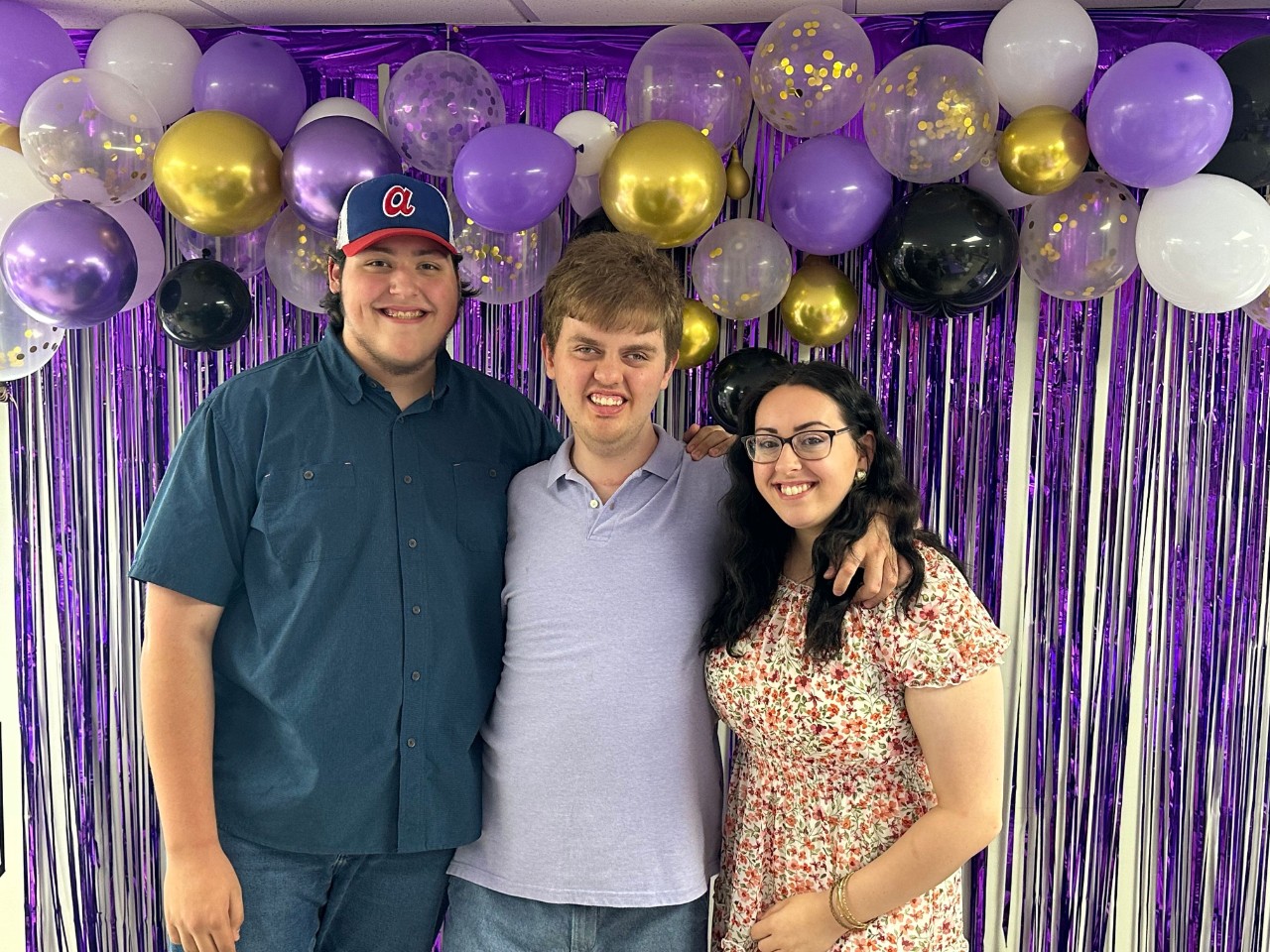 Harrison poses with Hannah and her younger brother, Noah Culwell. Hanna recently graduated from college and began her career as a teacher. Noah is under age 18 so not eligible to serve as paid staff through ECF CHOICES. Tecia says she has paid out of pocket for Noah to help out some as she searches for new support staff for Harrison. three white, young adults pose with their arms around each other in front of a purple drape background with purple, black, and gold balloons