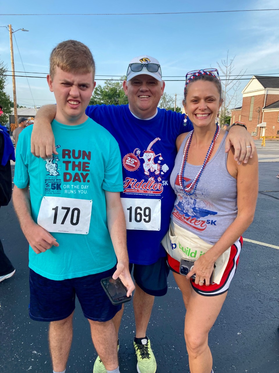 Harrison, dad John, and mom Tecia showed up big for the Smithville Fiddler 5K, which Tecia directs. A white young man and two older adults pose together, smiling at the camera. They are wearing race bibs and Fourth of July garb.
