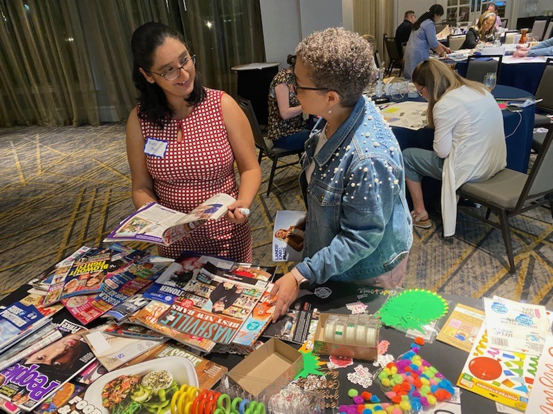Nidhi and Cat - 2023 Cathlyn stands at a table covered in craft supplies and old magazines, talking with a woman with dark hair and glasses who is holding an open magazine.