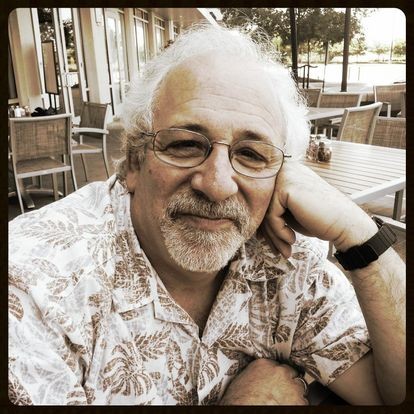 Ned Andrew Solomon A sepia-toned photo shows a white man with gray hair and mustache and goatee smiling at the camera, his head resting against his left hand. He is wearing a Hawaiian-style collared shirt, glasses, and a dark colored watch.