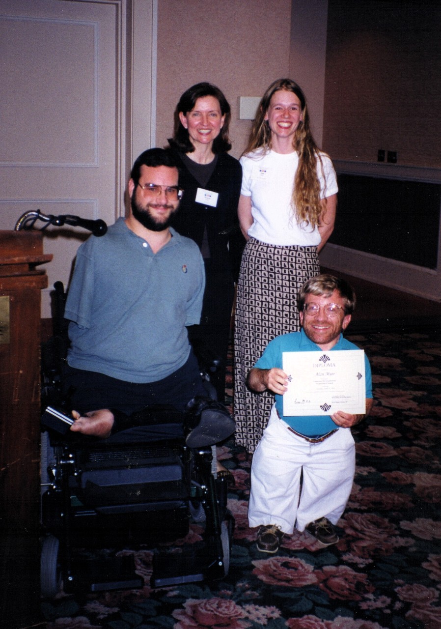 Partners 1999 Two white women, a white man who is a small person and is holding a certificate, and Kevin Wright, who is using a wheelchair and has limb differences, smile at the camera.