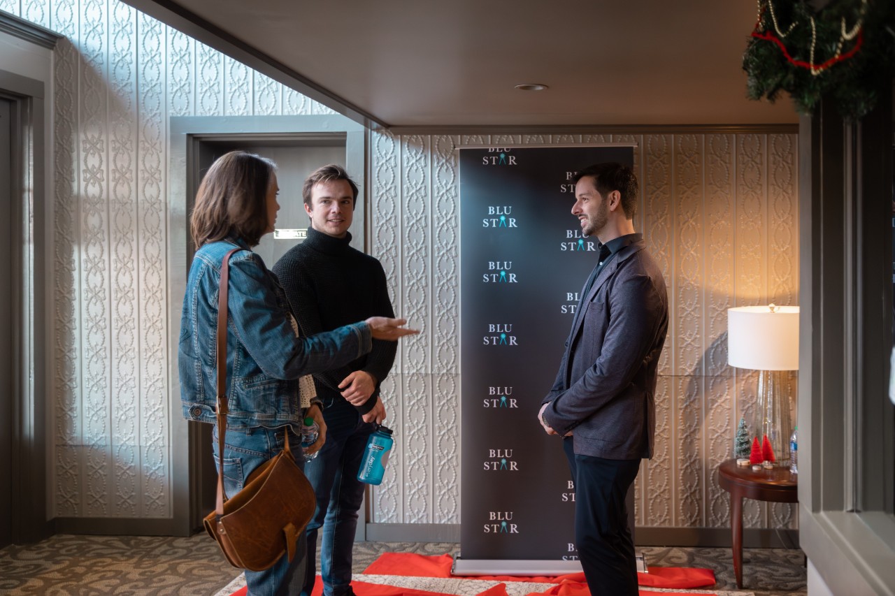 Alex chats with attendees at "Free Play" documentary premiere A young white man in a brown suit jacket and slacks listens as a couple of people talk to him, and all are standing in front of a photo backdrop with the Blu Star logo on it.