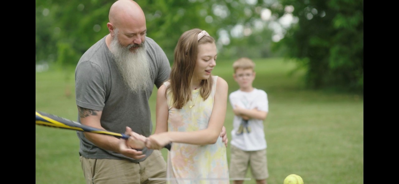 Still from "Free Play" a still shot from the documentary, showing an adult man with a gray t shirt and white beard standing behind a girl of about 10 or 11 helping her swing a baseball bat. the girl is in a white dress and has a visible disability. a young boy looks on from the background. they're all outdoors