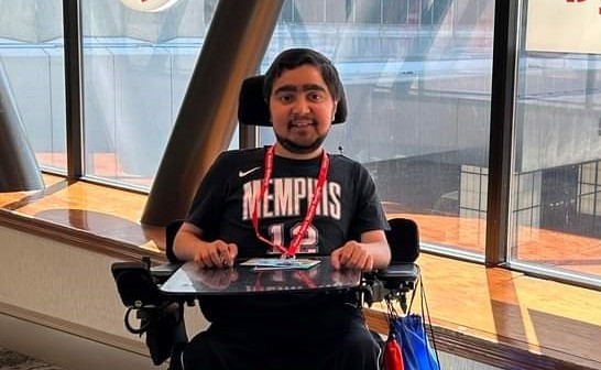 Ravi Lipman Image Description: Ravi Lipman, a person with medium-toned skin, sitting in a wheelchair and wearing a black t-shirt with the word "Memphis" on it. He is smiling and appears to be in an indoor setting with large windows behind him.