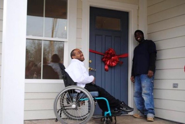 Image Description: Two men are in front of a blue door with a large red bow. One man is seated in a wheelchair, wearing a white shirt and black pants. He has light brown skin and is smiling. The other man is standing, wearing a dark shirt and blue jeans. He has dark skin and is also smiling. 