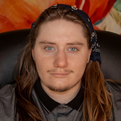 Image Description: A close-up portrait of a light-skinned person with long brown hair and blue eyes. He has a slight mustache and is wearing a dark-colored bandana with a pattern, tied around his head. He is also wearing a gray shirt with a black collar. The background is colorful, with warm tones and abstract shapes. The person is looking directly at the camera with a neutral expression.