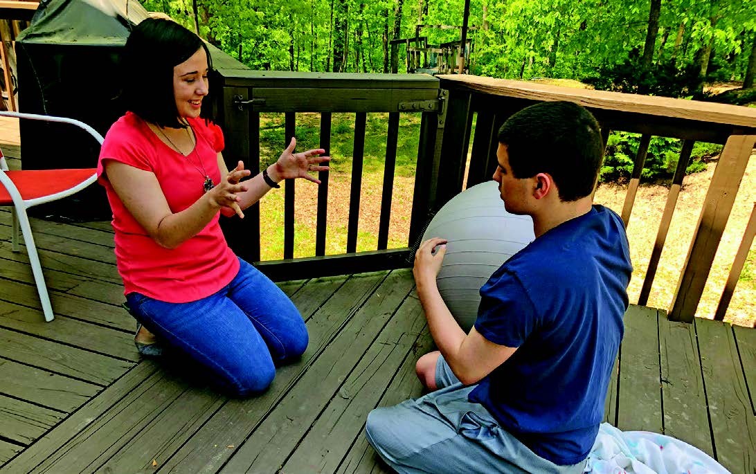 photo is of Lydia and Kevin, who are siblings, they are on their knees on the patio, passing a big therapy ball back and forth