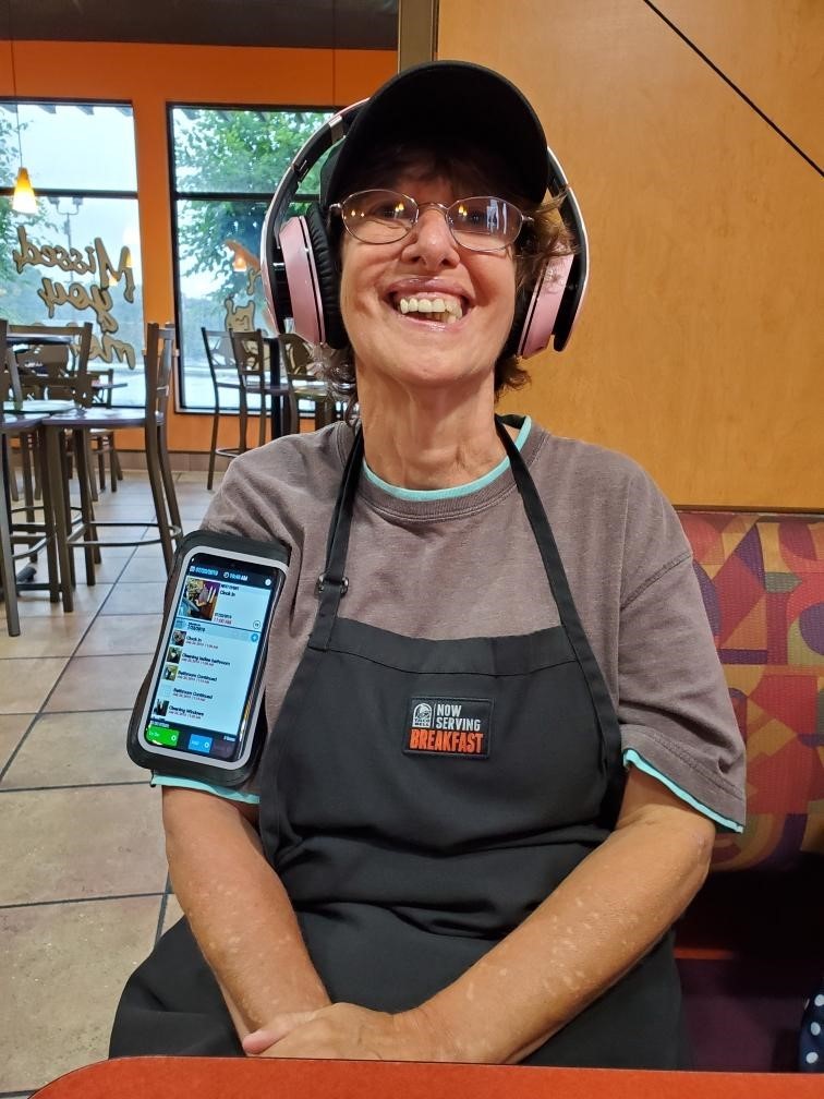 Anita at work a photo of Anita - a close-up of a woman working in a Taco Bell restaurant. She is wearing an apron over a gray sweatshirt. The apron has a badge with a Taco Bell company logo and the words, “now serving breakfast.” The employee also has a smart phone strapped to her right arm, and she is wearing large, pink headphones over her ears. She wears glasses, and has a huge smile. In the background you can see several customer tables with empty chairs, and the large, front windows