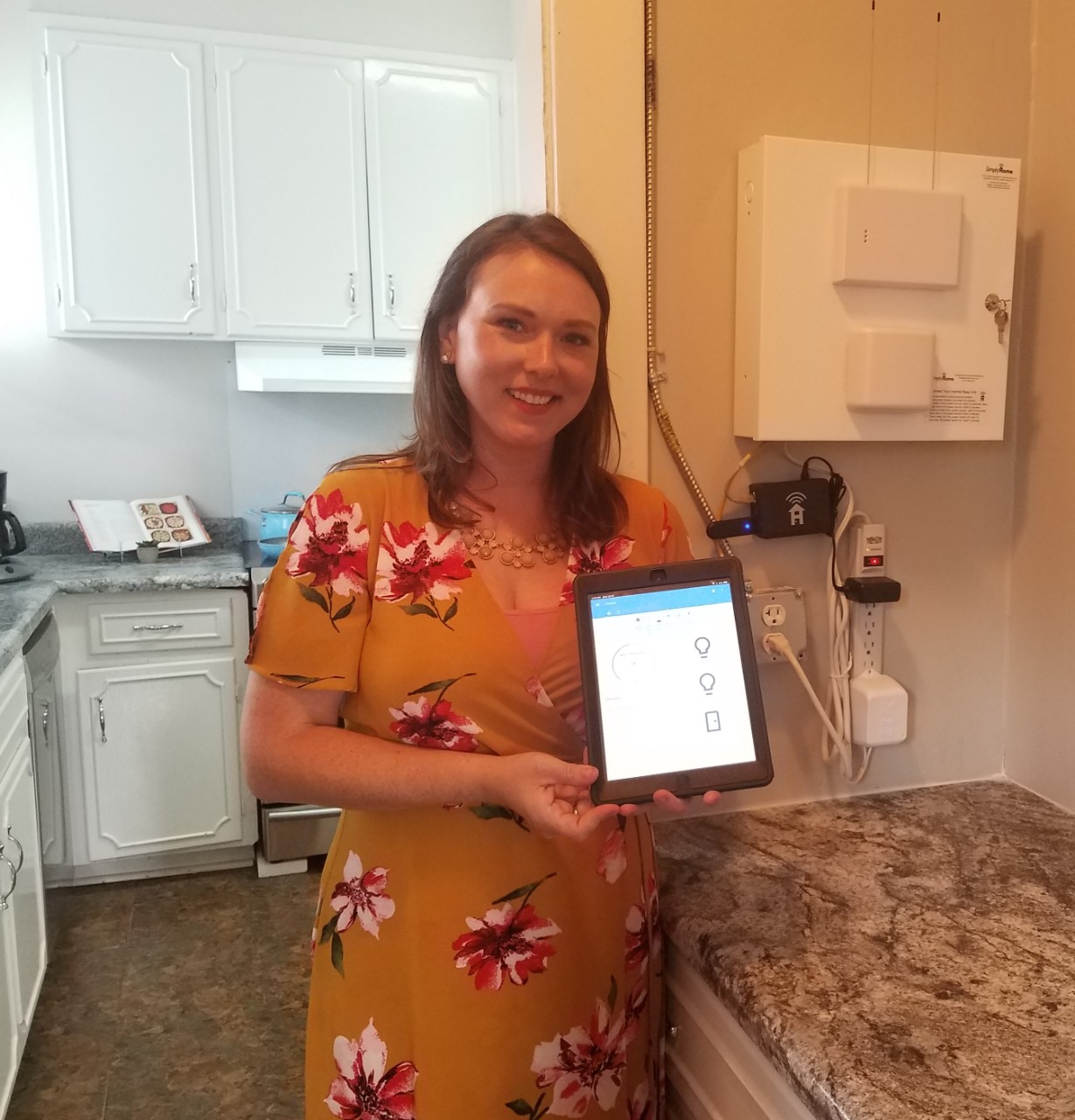 BG99 Enabling tech house Kate Kate Adams, a young woman in a gold floral dress, stands in the kitchen of the technology model home described in this article and holds up an ipad showing an open app