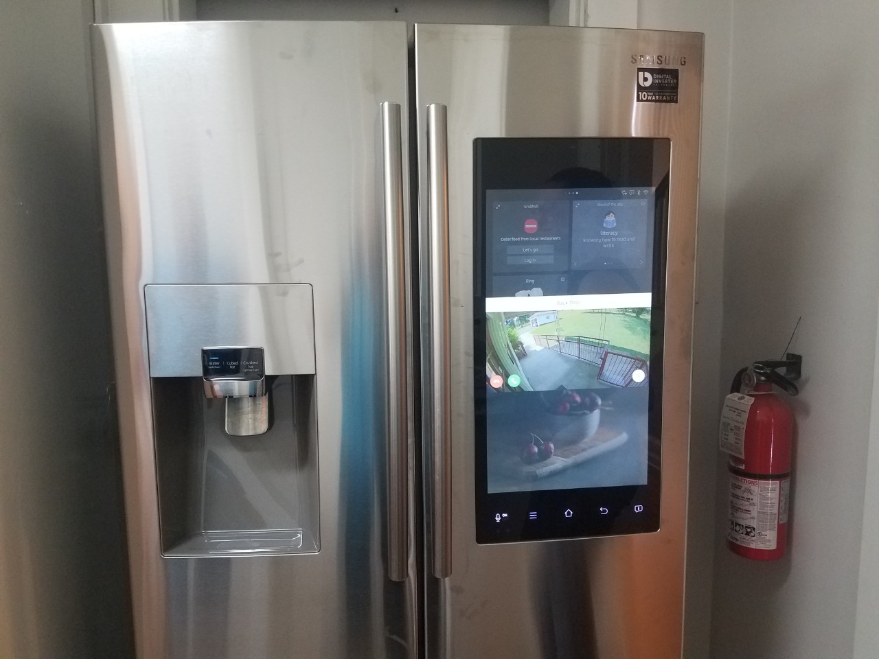 "Smart" refrigerator a photo of a stainless steel refrigerator with a large digital panel in one of the doors that shows the view outside the front door from a camera, and has other features where a person can make shopping lists, use a calendar and many many other tech functions
