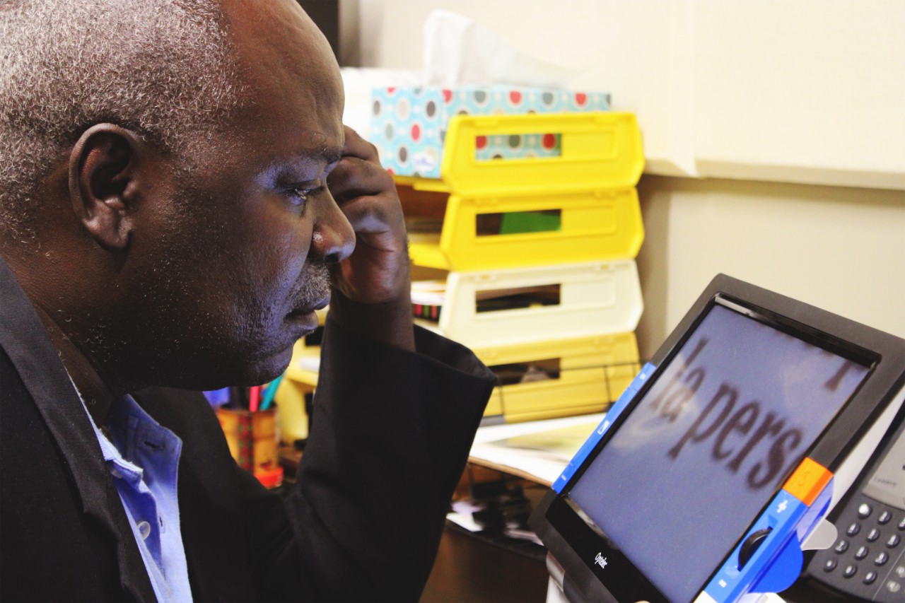 An African gentleman with white hair and a black suit jacket looks at a CCTV with enlarged text at a desk