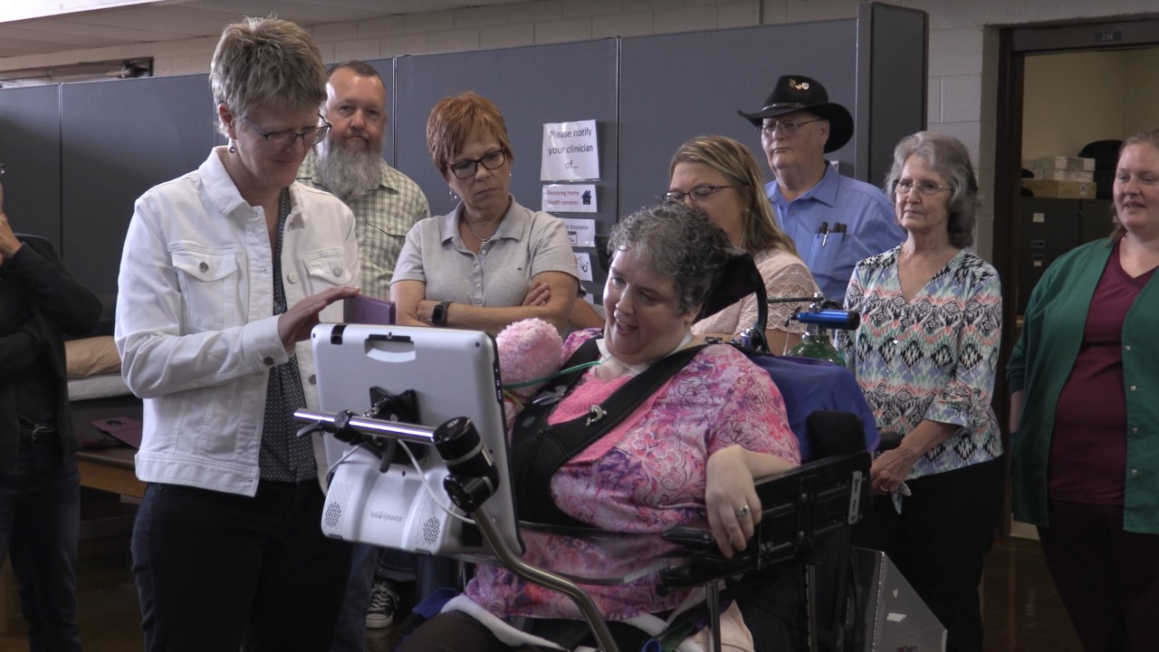 Technology innovations allow people with limited movement and speech to drive wheelchairs with eye movements and communicate with family, friends, and staff A group of a few adults stand around a middle aged gray haired woman in a power wheelchair who is using a tablet to communicate with them with a text to speech app