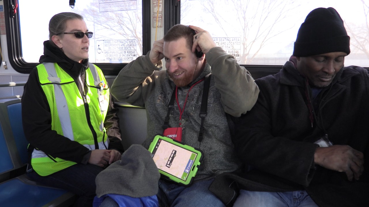 Kenny uses technology on his iPad to help him use public transportation and be more independent. A young man with an intellectual disability is pictured seated on a bus, looking excited, as he puts earphones on and he has a tablet in his lap, attached with a lanyard around his neck to help him communicate
